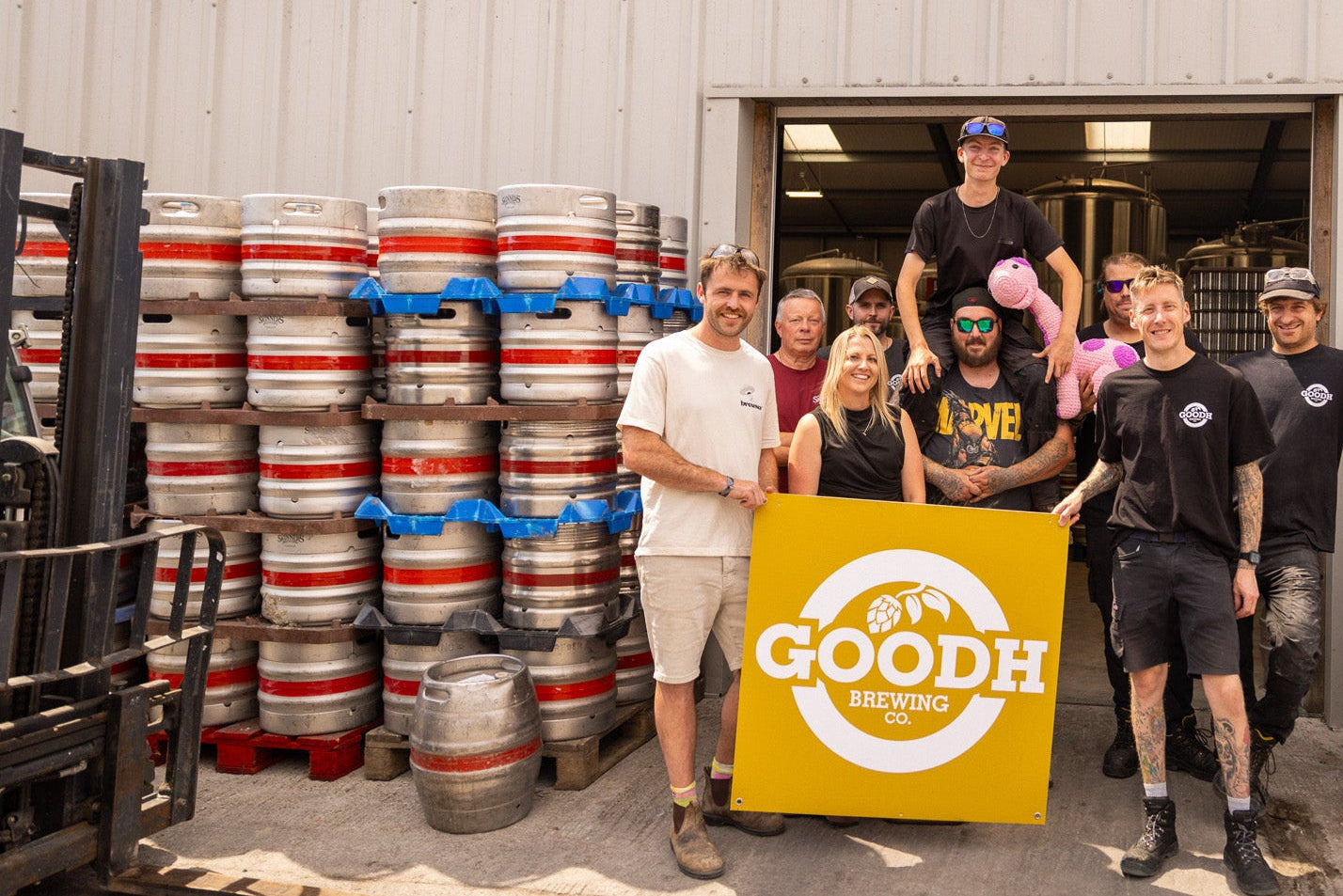 A group of people standing in front of a warehouse doorway with stacks of metal beer kegs on both sides, holding a large yellow sign that reads 'GOODH BEER TASTING'.