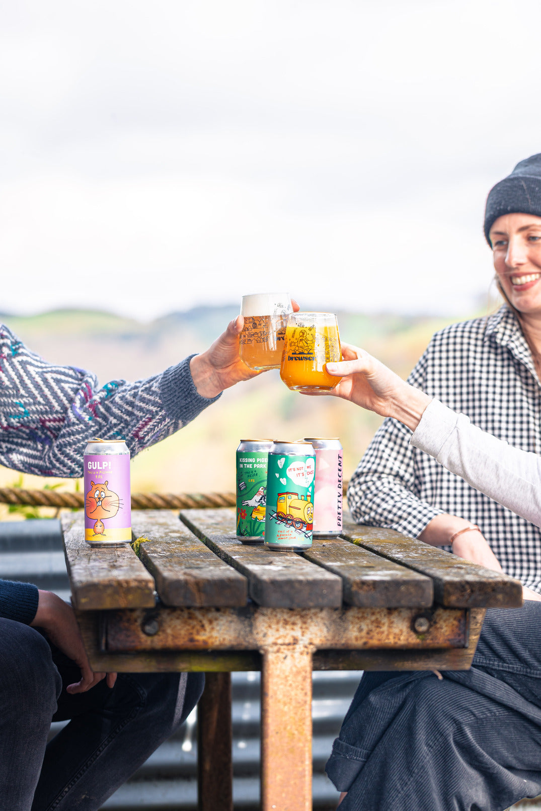 Two people toasting with drinks at a wooden table outdoors.