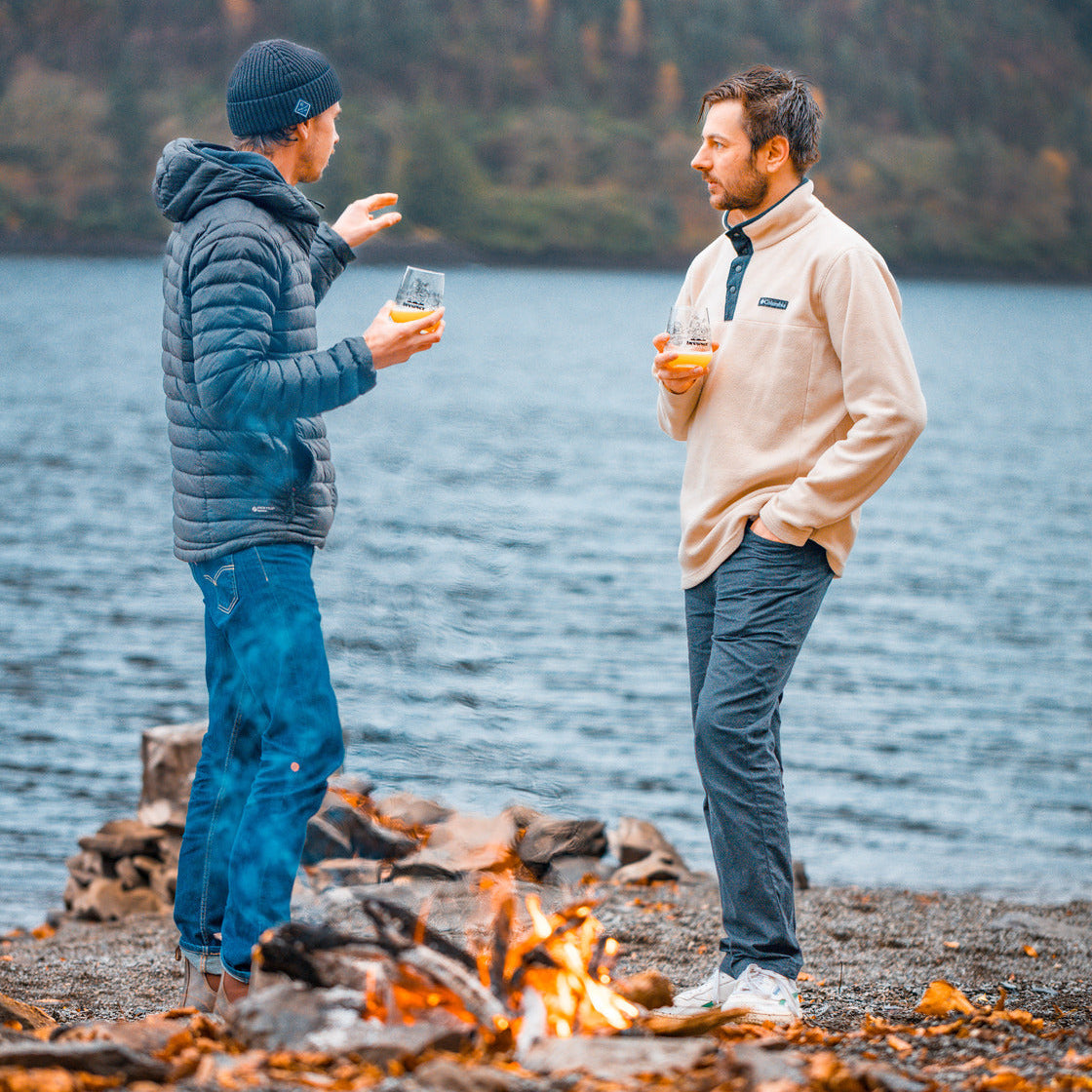Two people by a campfire by a lake, enjoying drinks.
