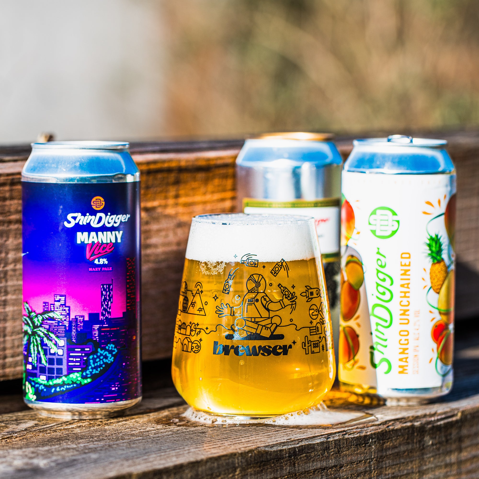 ShinDigger Manny Vice Hazy Pale and Mango Unchained cans with a Brewser glass on a wooden surface