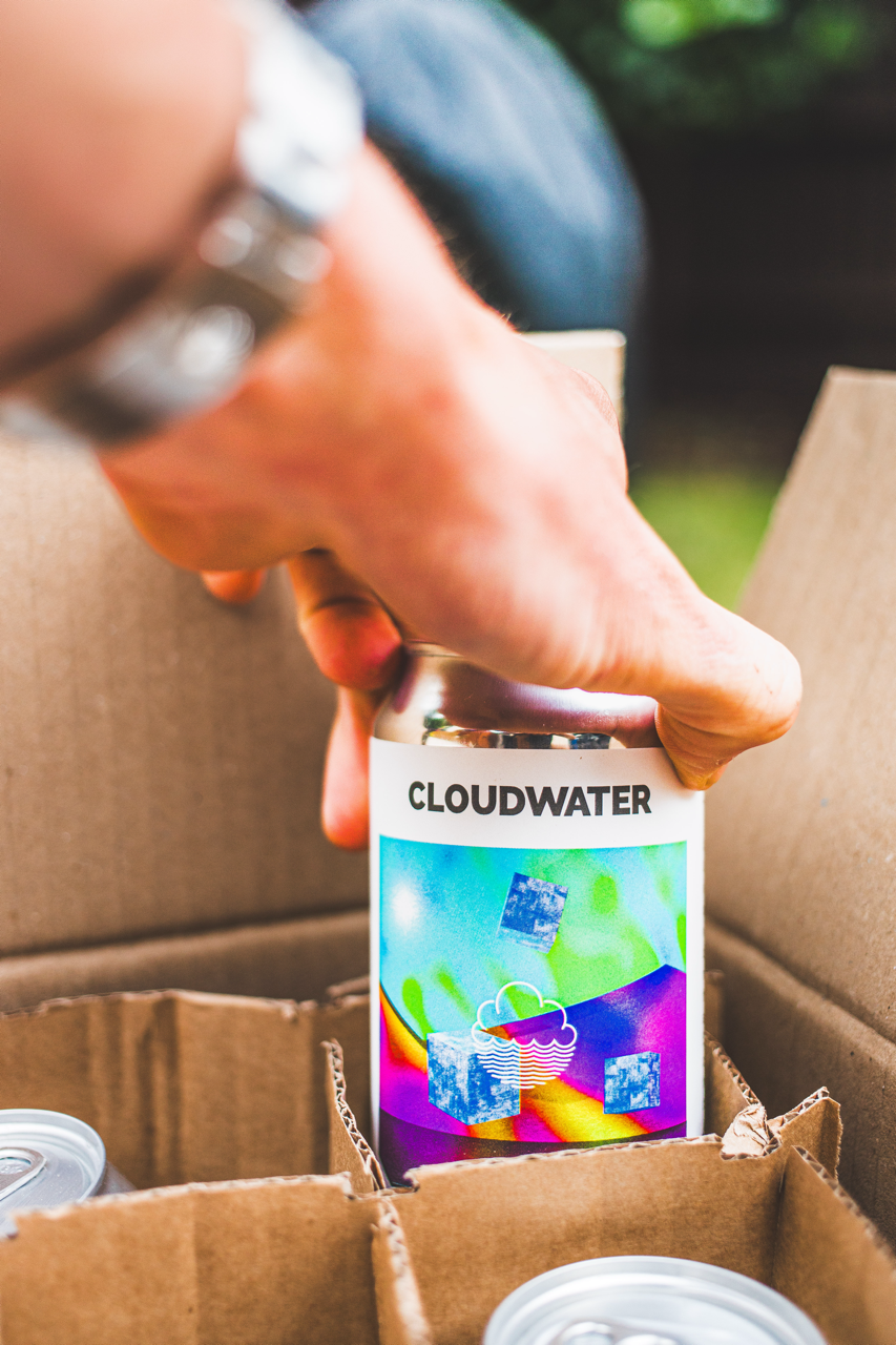 Hand holding a Cloudwater can over an open Brewser subscription box of craft beer cans