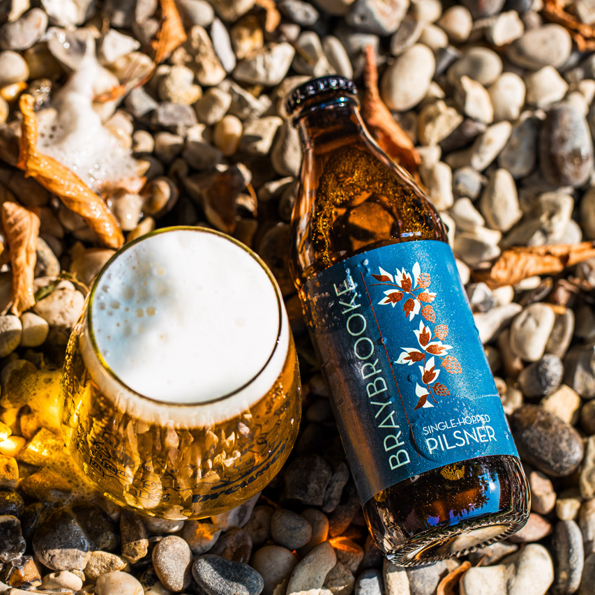 Braybrooke Single-Hopped Pilsner bottle and a poured pint on pebbles, Brewser craft beer subscription