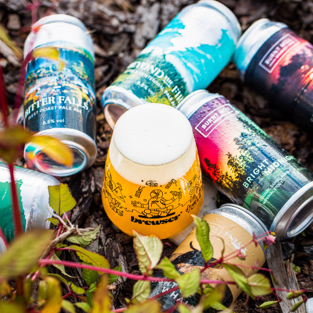 Brewser glass with Burnt Mill Brewery and Finback craft beer cans on natural foliage including Bitter Falls West Coast Pale, Beyond the Firs and Bright Midnight - Brewser craft beer subscription direct from brewery UK