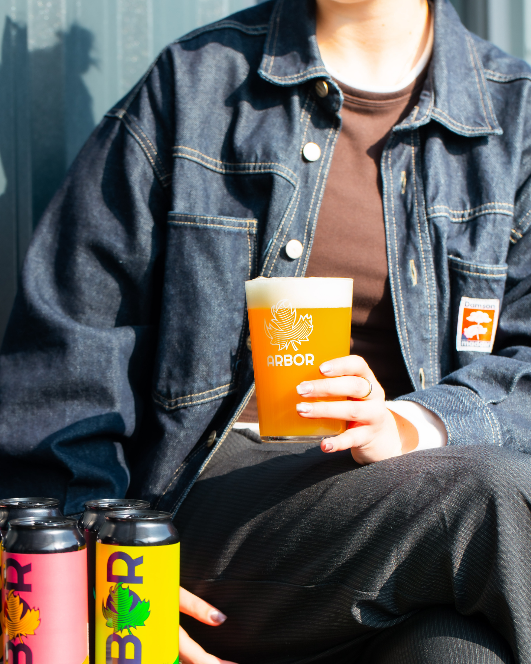 Person holding a glass of Arbor Brewing beer with multiple cans in front