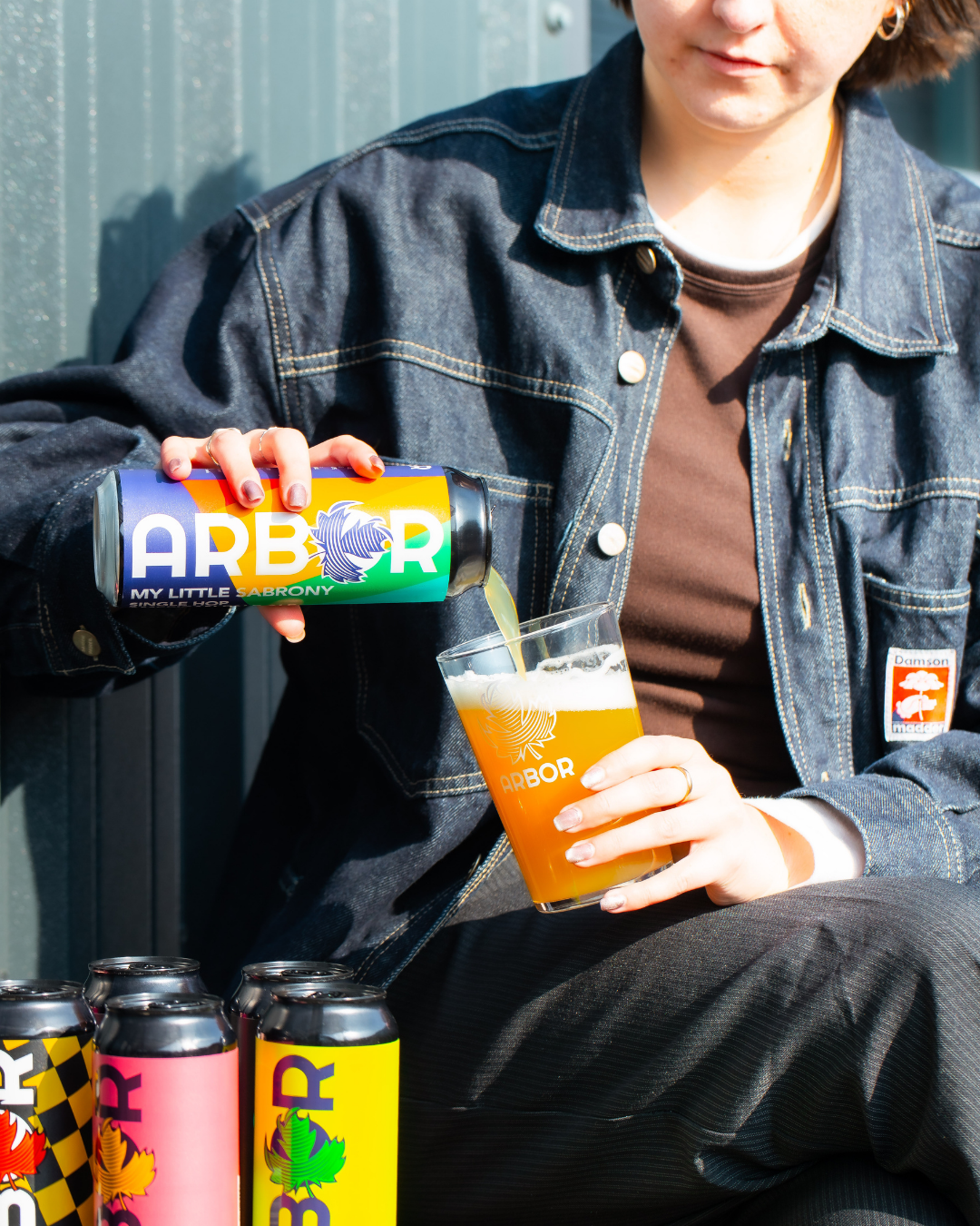 Person holding a can of Arbor Brewing beer and a glass of beer against a metal wall.