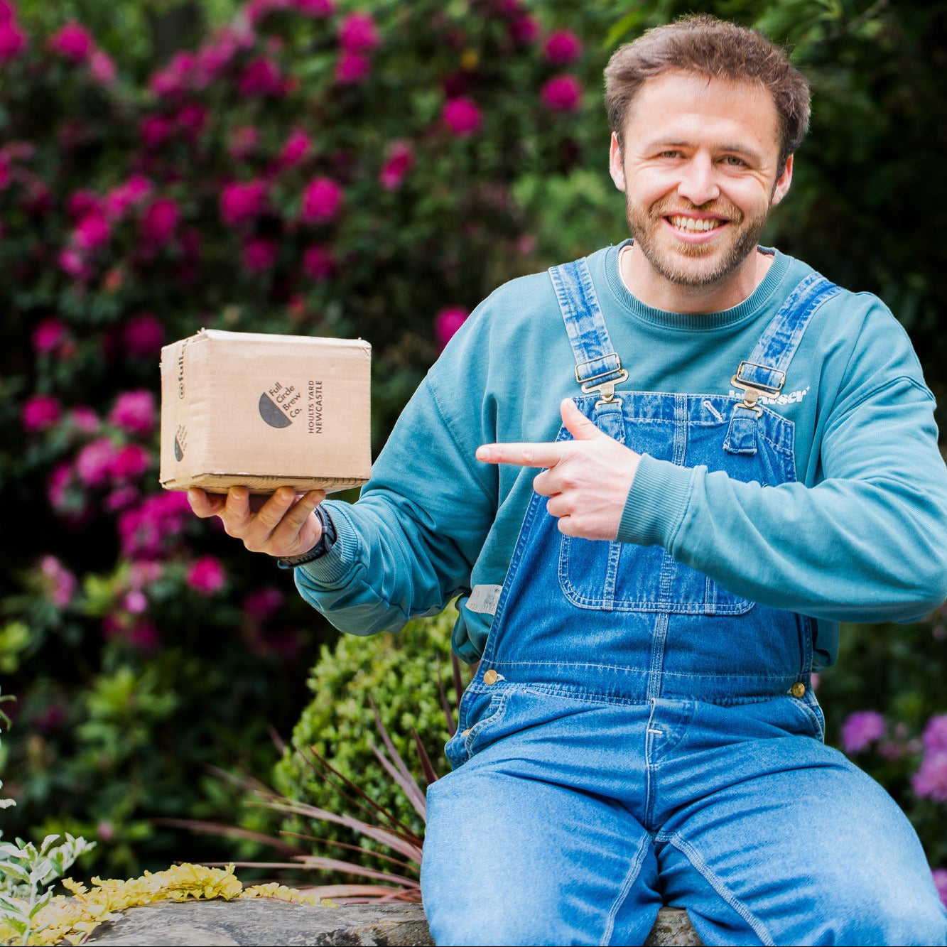 Happy man in blue overalls holding and pointing to a Brewser craft beer subscription box outdoors surrounded by flowers - buy craft beer online direct from independent brewery with Brewser UK