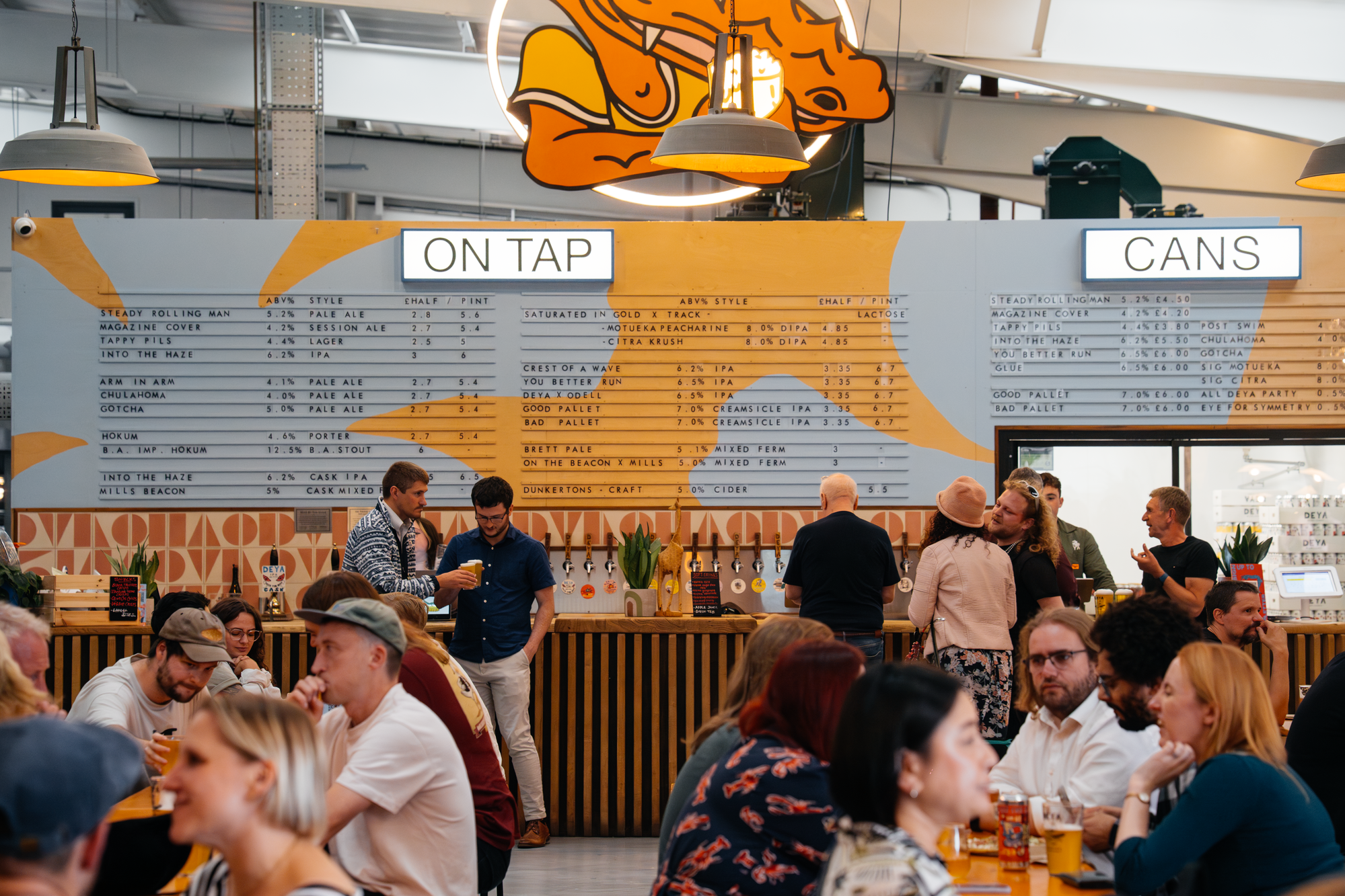 Busy craft beer tap room with people enjoying cask and keg ales, On Tap and Cans menu boards visible - craft beer community event supported by Brewser craft beer subscription UK