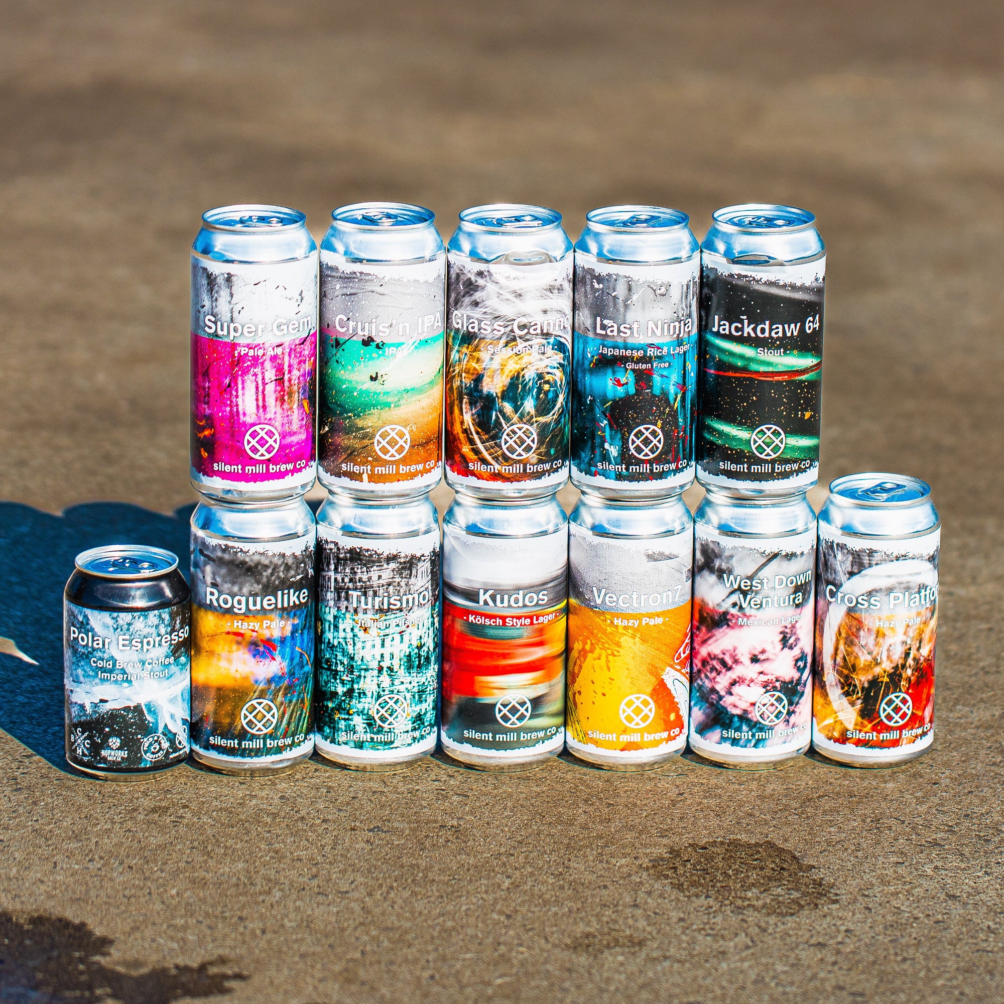 Full Silent Mill Brew Co craft beer range including Roguelike Hazy Pale, Jackdaw Stout, Kudos Kolsch, Cross Platform and more cans on sandy surface - Brewser craft beer subscription direct from brewery UK